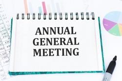 AGM Annual General Meeting is written in red on a white piece of paper on a light yellow background next to a laptop, pen, magnifying glass, glasses and a green plant.