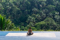 The girl testifies sitting on the rug in the lotus position on the background of the tropical jungle. Back view
