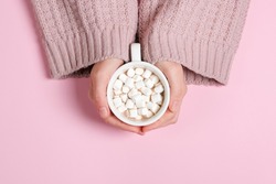 Girls hands in powder-colored sweater holding large cup of cocoa or hot chocolate isolated on pink background.