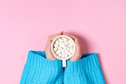 Top view on Girls hands in blue cozy oversized sweater holding large cup of cocoa or hot chocolate isolated on pink background.