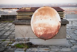Old rusty satelite antenna dish on roof of the building hangs on the rooftop chimney ventilation system. Parabola satellite receiver. Outdated corroded equipment.