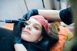 Closeup of young woman's face with tatoo artist's hands holding tatoo machine upon her brow. Permanent make up, tatoo and brow making professional sphere.