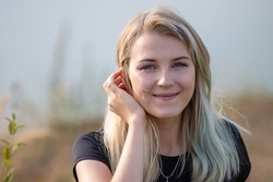 Close-up of a gentle smiling girl with  gray eyes. She has blonde hair and a black T-shirt. Horizontal photo of a cute girl 20 years old on a blurred background of nature.