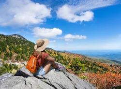 Hiker girl sitting on a cliff edge enjoying scenic fall view. Woman relaxing on top of autumn mountain Grandfather Mountain State Park, Banner Elk, North Carolina, USA.