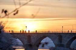 Selected focus. People walking on a stone bridge at sunset and the frozen trees in winter at the golden hour. Stone arches, reflection of the sun on the river. Two bridges. Horizontal view