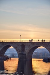 People walking on a stone bridge at sunset and the frozen trees in winter at the golden hour. Stone arches, reflection of the sun on the river. Two bridges. Sewers in the background. Vertical view