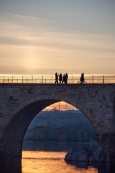 People walking on a stone bridge at sunset and the frozen trees in winter at the golden hour. Stone arches, reflection of the sun on the river. Sewers in the background. Vertical view