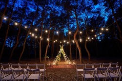Festive string lights illumination on boho tipi arch decor on outdoor wedding ceremony venue in pine forest at night. Vintage string lights bulb garlands shining above chairs at summer rural wedding.