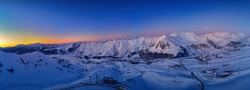 Wide aerial panorama of snowy mountain ridge on winter sunrise. Stunning mountains range covered with snow powder on ski resort at sunset. Moon above caucasus peaks skyline in the dusk at night.