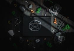 Black leather book with magical white symbol,stones,black candles,green moss,dried flowers, ritual sticks,tarot cards,wife paper lie on a black wooden background,flat lay .Esoteric concept,dark style.