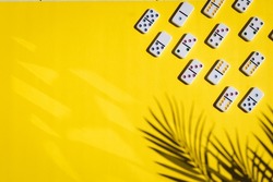 White dominoes with colorful dots lie on the right on a yellow background with a shadow of a palm tree branch with copy space and sun glare on the left, flat lay close-up. Concept summer board game.