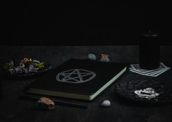 BlaA black leather book with a magical white symbol, black candles, black metal saucers, dried flowers, ritual sticks, tarot cards, a pen and stones lie on a black wooden background, close-up side vie