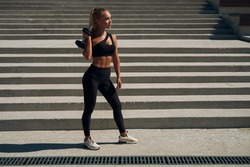 Athletic blonde holds a dumbbell on her shoulder. young woman in black suit trains arms at sunrise.