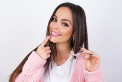 young beautiful caucasian woman wearing pink knitted jacket over white wall holding an invisible aligner and pointing to her perfect straight teeth. Dental healthcare and confidence concept.