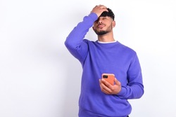 Upset depressed young arab man with curly hair wearing purple sweatshirt over white background makes face palm as forgot about something important holds mobile phone expresses sorrow and regret blames