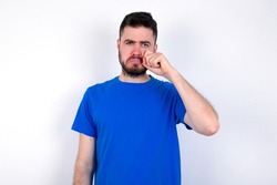 Disappointed dejected Young caucasian man wearing blue T-shirt over white background wipes tears stands stressed with gloomy expression. Negative emotion