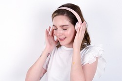 Pleased young caucasian woman wearing white T-shirt over white background enjoys listening pleasant melody keeps hands on stereo headphones closes eyes. Spending free time with music