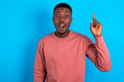young handsome man wearing pink sweater over blue background pointing finger up and looking inspired by genius thought, showing good idea sign, having clever solution in mind
