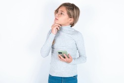 beautiful caucasian blond teen girl wearing gray turtleneck sweater over white wall thinks deeply about something, uses modern mobile phone, tries to made up good message, keeps index finger near lips