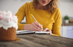Low angle of joyful woman sitting at kitchen table and making notes in her notebook with pencil. Glazed easter cake is standing in front of her. Focus on female