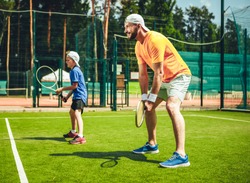 Full length side view beaming bearded male and outgoing boy situating in special pose while having fun during game with rackets in hands. They standing on court