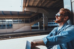 Pleasant smiling young man sitting while resting on the railway station and waiting for a tain