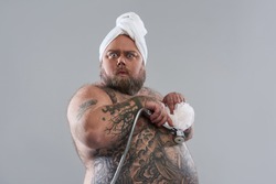 Waist up of funny surprised fat man standing isolated on the grey background and pressing shower head with shower puff to his tattooed belly