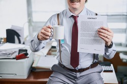 Mature old fashioned businessman holding lucrative contract and a cup of tea in hands with his office desk on background