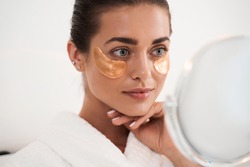 Under eye masks for puffiness, wrinkles, dark circles. Eye patches concept. Satisfied woman in white bathrobe applying gold eye patches in bathroom while looking at the mirror