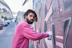 Handsome multicultural street artist with dreadlocks standing near the wall while painting colorful graffiti at the street. Urban, street art, millennials generation, mural concept