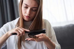 Perturb pretty woman looking at damaged hair tips and worries about split hair, while brushing her hair at home. Stock photo