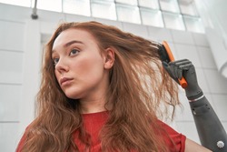 Perturb pretty woman with hand implant looking at damaged hair tips and worries about split hair, while brushing her ginger hair at home. Stock photo