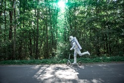 Spaceman wearing white armor is using scooter and driving through roar among trees. Profile. Copy space on left side