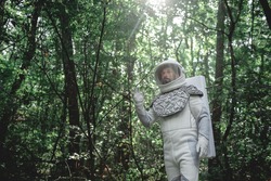 Surprised cosmonaut wearing white armor and helmet is standing among trees. He looking ahead with bewilderment in his eyes. Low angle. Copy space on left side