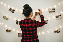 Young beautiful woman hangs a photos on a garland with light on the wall. Bokeh, girl in red checkered shirt, comfort at home, New Year's mood, Christmas decorations.