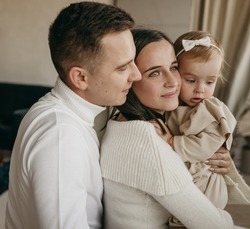 The portrait of beautiful family of mother, father, daughter in white clothes near mirror at home. Nice modern minimal interior. Beige colours. Happy people, studio photoshoot, hugging.