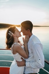 The portrait of cute loving couple hugging in the white boat in lake at sunset. Summer vacation, love, kissing, honeymoon. Beautiful view, warm sunlight, blue water of sea. emotional man and woman.