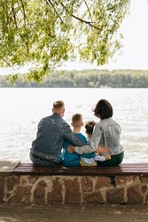 The beautiful family of four people, father, mother and two sons sitting at bench near lake. Happy family, walking outdoors. Childhood, lovely parents. Hug from behind.
