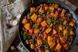 Homemade stew with ground beef, pumpkin and kidney beans cooked with vegetables such as tomato, bell peppers, garlic and herbs. Served in rustic cast iron skillet on wooden table. Overhead view.