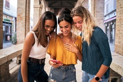 Three happy women using mobile phone outdoors. Group of smiling female friends watching social media at smartphone