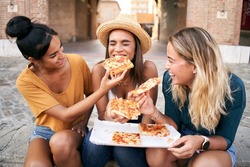 Happy girlfriends eating pizza street food at the city. Female tourist group of three woman having fun. One girl feeds the other. Lifestyle and friendship concept