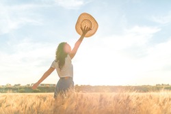 Young woman in a ripe wheat field celebrating with a hat in hand in a sunny wheat field. Concept of prosperity and warmth.