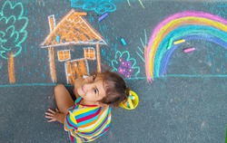 The child draws a house and a rainbow on the asphalt with chalk. Selective focus. Kids.