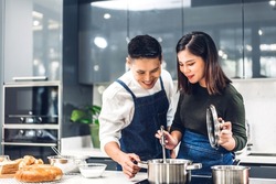 Young asian family couple having fun standing near stove and cooking together.Happy couple looking and smelling tasting fresh delicious from soup in a pot with steam at white interior kitchen