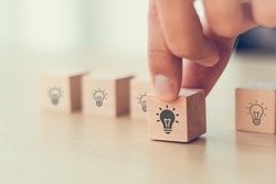  Idea and creativity concept. Male hand holds wooden cube with light bulbs icon on light background. Teamwork brainstorming ideas, evaluation and selection for business development. Banner, copy space