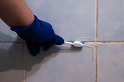 A hand in a blue glove cleans the dirty joints between the tiles in the bathroom with an old toothbrush. Toxic black mold in the bathroom during the cleaning process.
