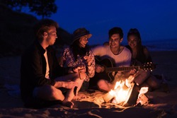 young and cheerful friends sitting on beach and fry sasuages or weenies in bonfire One man is playing guitar. Music on Wild beach. Happy couple relationships.