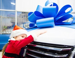 Little girl in red Santa Claus outfit hugs the car in front. Auto as a present with a big blue bow. The child's face is not visible
