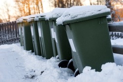 Green garbage cans in a row with snow on top on cold winter day with orange sunlight in background
