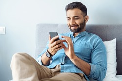 Smiling young adult indian freelance business man using mobile phone checking social media network feed or message chat sitting on bed at home. Online digital communication, rest after hard work day
